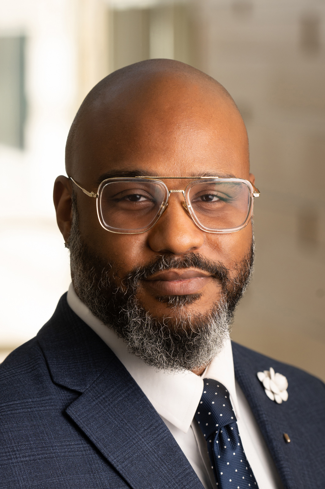 A man standing with eyeglasses, shaved head, a beaded chain and a beard smiling