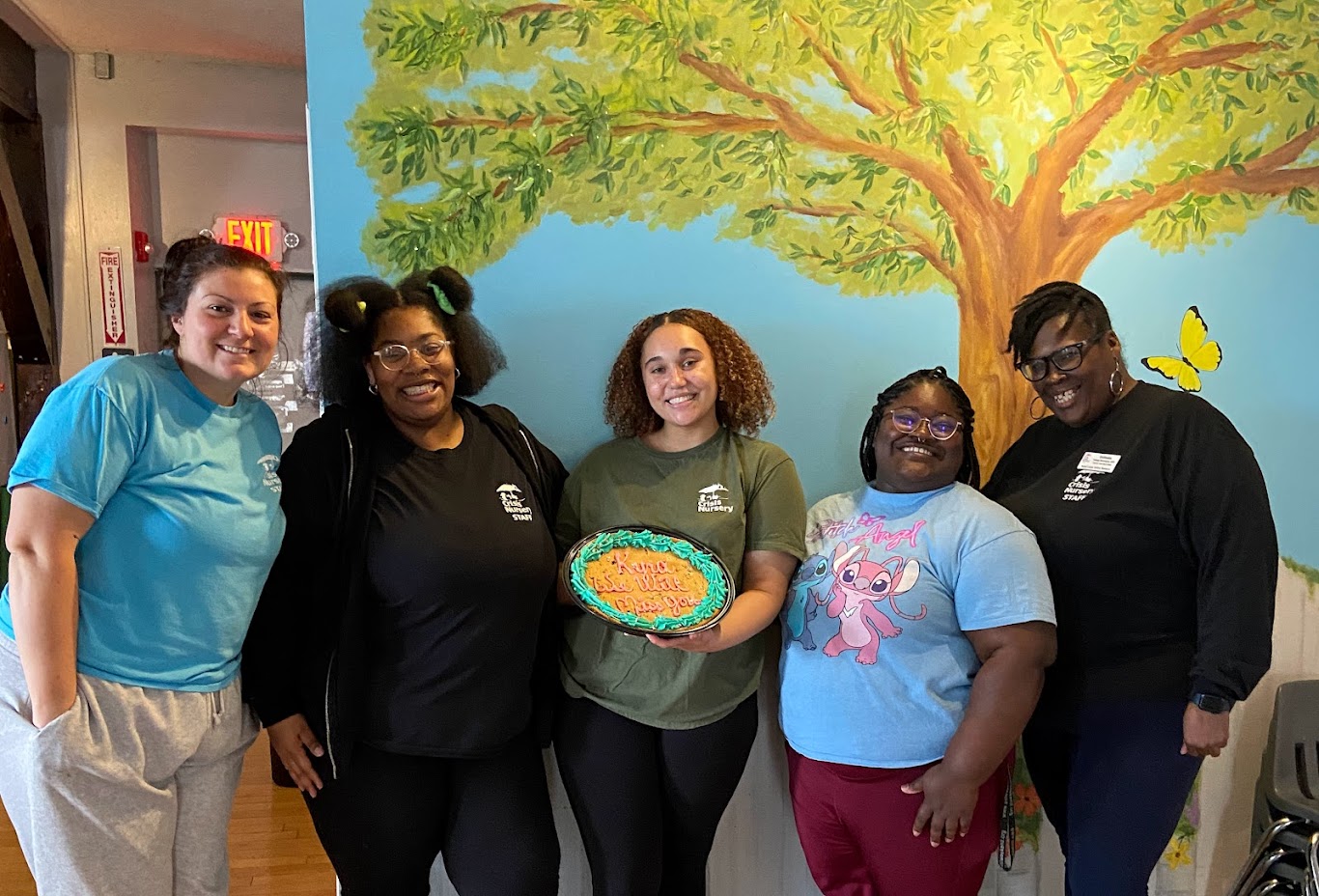 Five people standing in a classroom, one holding a cookie cake, with a mural of a tree behind them