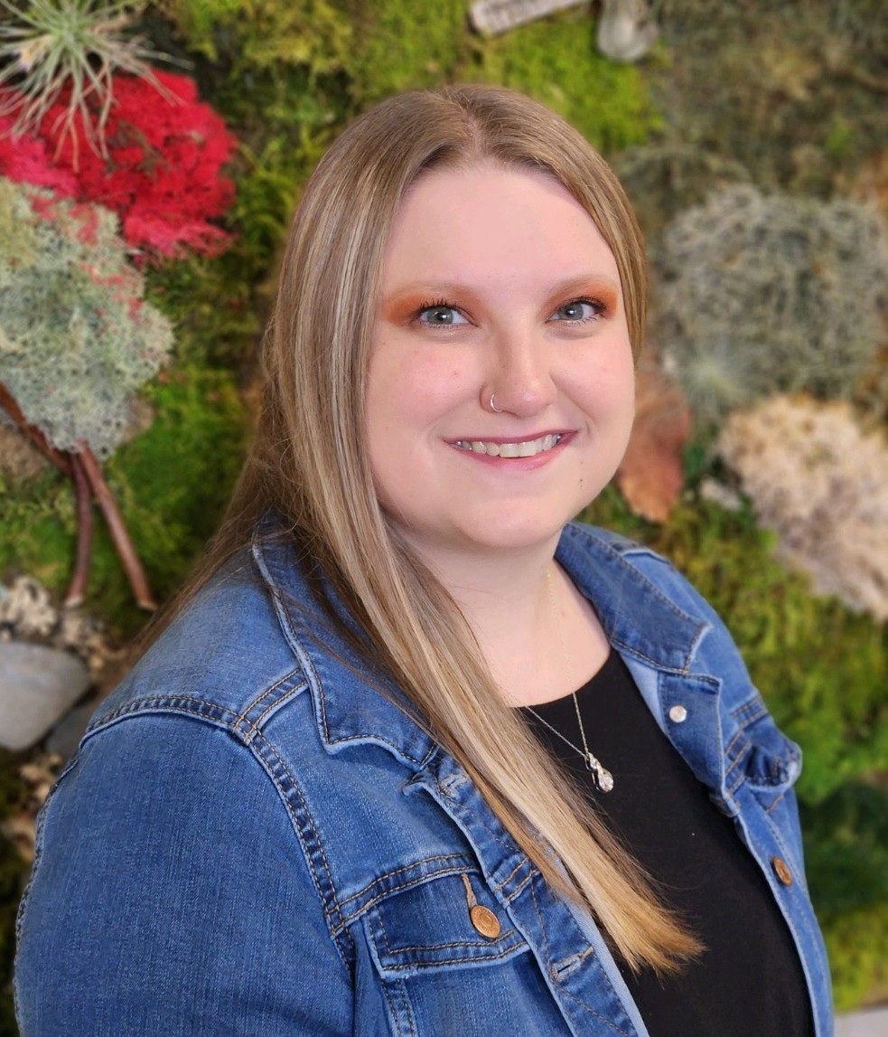 The image shows a person with long, straight blonde hair, smiling and looking towards the camera. They are wearing a denim jacket over a black top, with a pendant necklace visible. The background consists of a textured arrangement of greenery and moss, interspersed with clusters of bright red and muted gray foliage, providing a natural and vibrant backdrop.