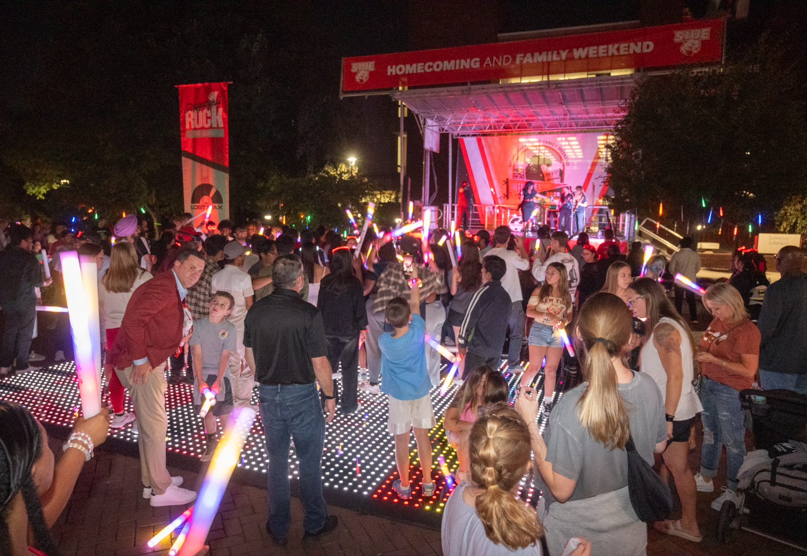 A crowd dances with colorful glow sticks on a lighted floor as live music plays beneath the Homecoming &amp; Family Weekend banner at Cougarfest.