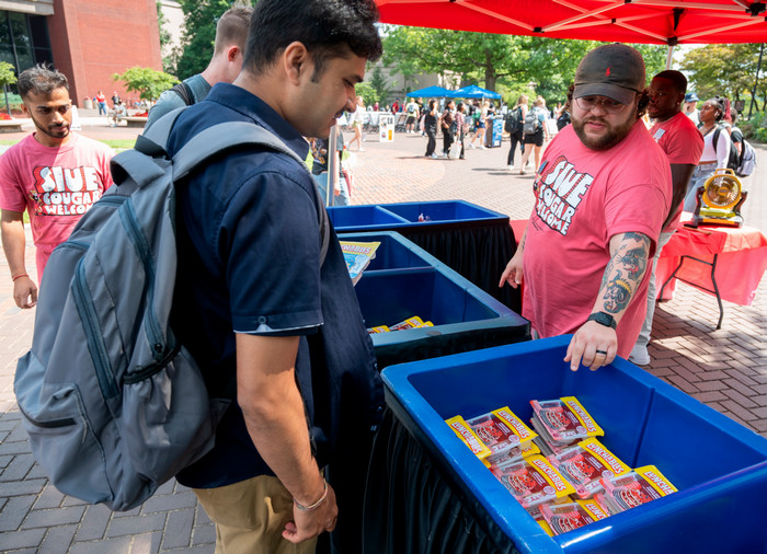 Two students browsing nostalgic Lunchables foods in offering bins on the quad. Two SIUE staff members wearing "SIUE Cougar Welcome" tshirts are pointing and assisting them.