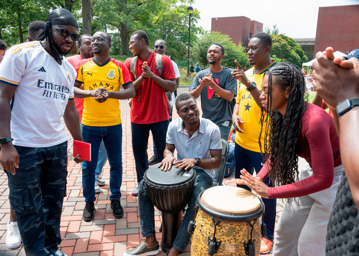 Group of seven international students playing large drums on the quad, clapping and smiling