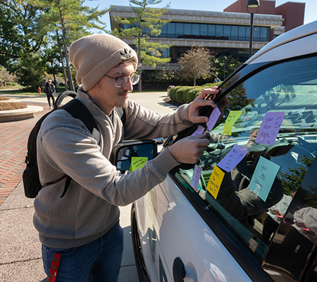 student placing a sticky note on a police SUV window outside of the MUC on a sunny fall day and rendleman hall is behind him