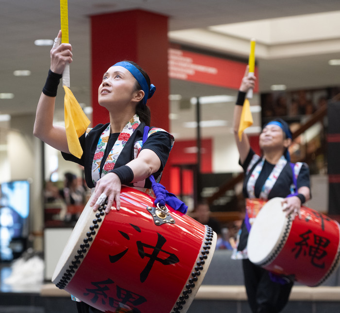 Two taiko drummers performing each with a single taiko in traditional wear in the goshen lounge while the closest one holds a drumming tool into the air proudly