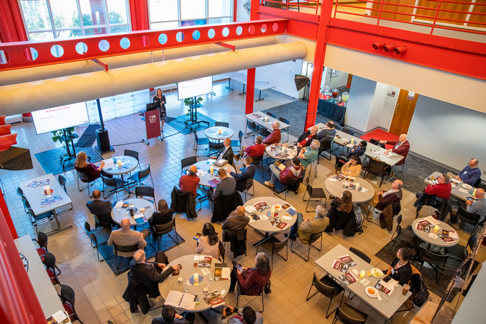 Photo of Keynote speaker Kelli delivering an address to a sunlit atrium with listeners gathered around circular tables