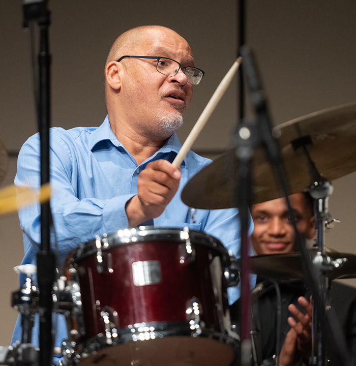 Morrison on drums, He is about to strike a cymbal and a student behind him admires his technique