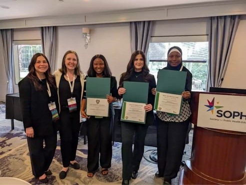 three students Glory Omole, Abigale Kambarian, and Maryam Musa with certificate awards, being honored by two women members of the Society for Public Health Education in a conference room