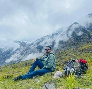 Kridip Amgai in Nepal. He is resting on a mountain side with lush green grasses, heavy cloud cover over mountains in the near background. A hiking backpack is beside him.