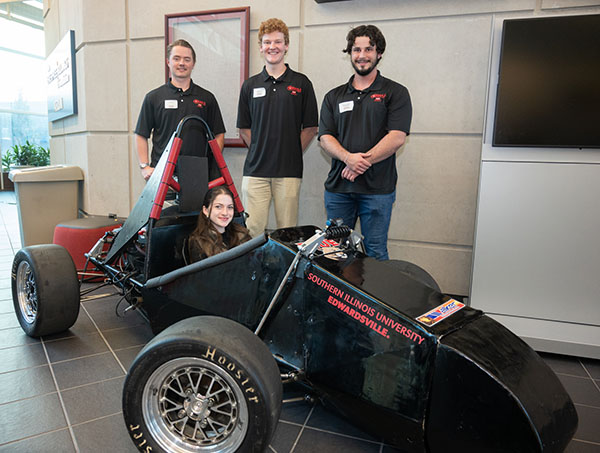 SIUE Formula SAE Vehicle in the Engineering Atrium with four members of the Formula SAE Team. The Tires of the vehicle are extra large and the cockpit of the vehicle is wedge shaped and a student on the team is sitting inside as if to drive