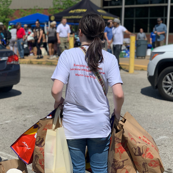 Photograph of back of volunteer in white tee with red lettering carrying bags of donations and supplies