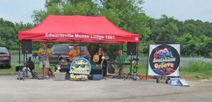 photo of the fundraising tent outside the Moose lodge on a sunny day. There is a band playing and a pond behind them,