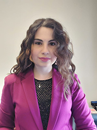 Portrait of Milena wearing a pink blazer inside an office. Her hair is wavy and she is wearing a necklace over a black top.