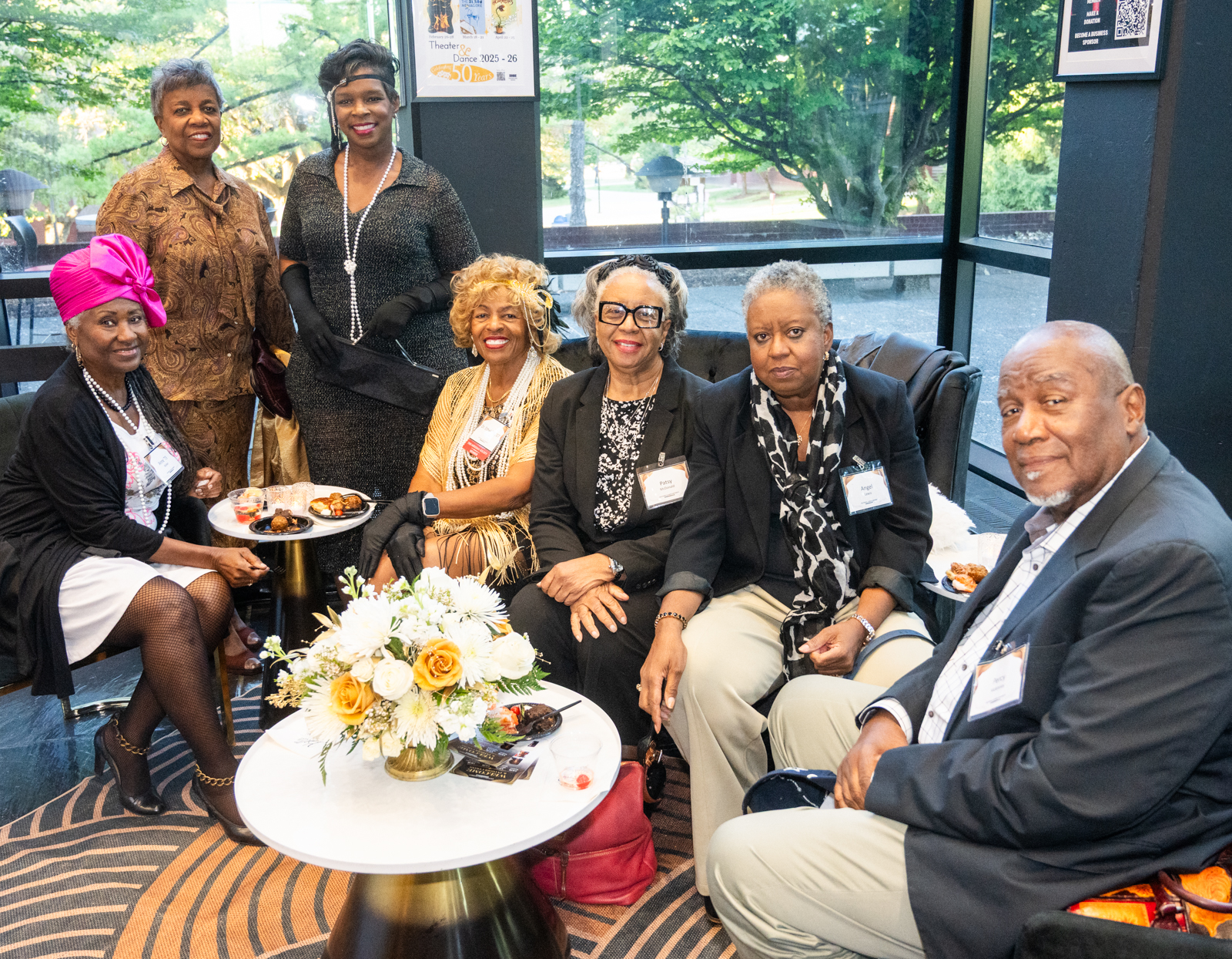 Group of attendees, including President Ethel Shanklin, seated and enjoying refreshments on a gold rug and cocktail table with gold flowers. They are dressed like 1920's aristocrats with pearls and hair fascinators, in front of a Dunham Hall window 