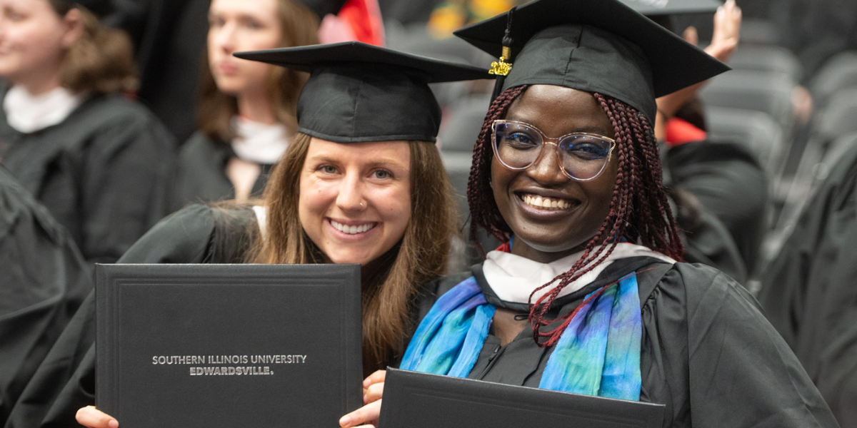 Two graduate students in regalia holding up a black SIUE diploma and smiling at the camera