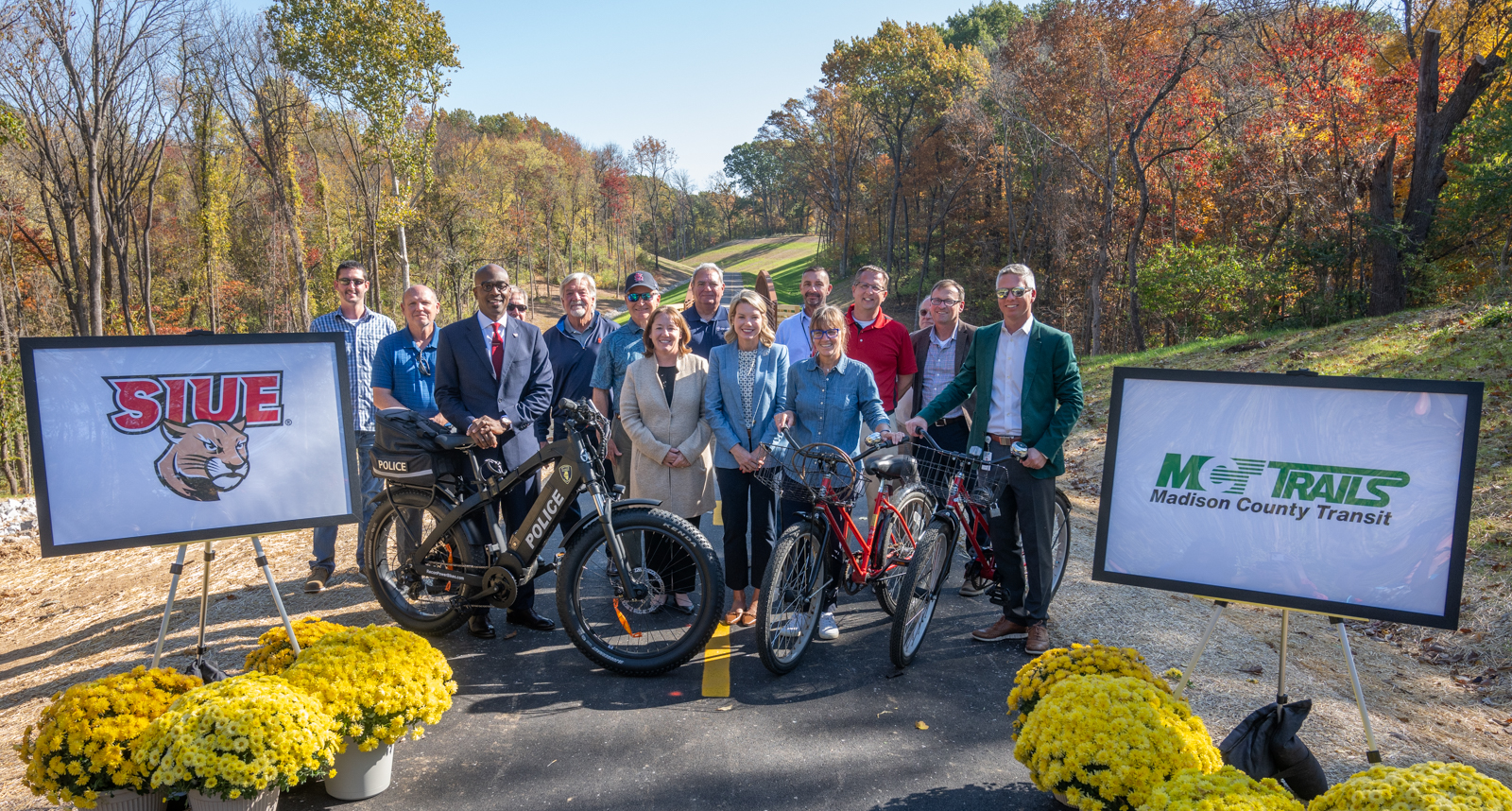 Group of people standing with bicycles on a newly completed trail surrounded by fall foliage, with SIUE and Madison County Transit MCT Trails signs displayed on easels.