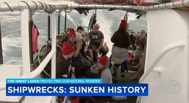 Photo of about ten people seated in the back of a small vessel cutting through water on a cloudy day. They are wearing red watch caps and appear excited. In the foreground, text reads "Shipwrecks: Sunken History"