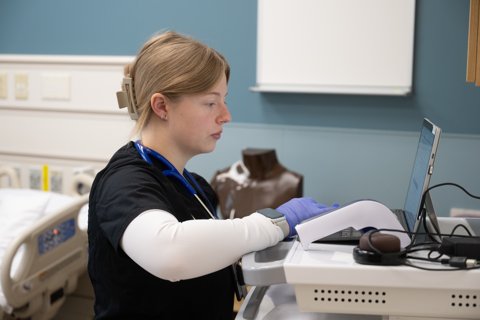 A person in medical scrubs and gloves works at a computer station in a clinical simulation room, with a stethoscope around their neck and a medical manikin visible in the background.