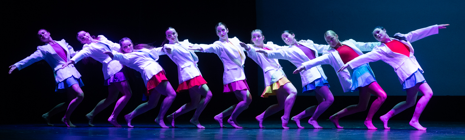 Ten dancers arranged in a chorus line formation on Dunham Hall Stage. They are wearing white work blazers over their brightly colored costumes, and they are leaning back symmetrically with an accordion or domino effect. 