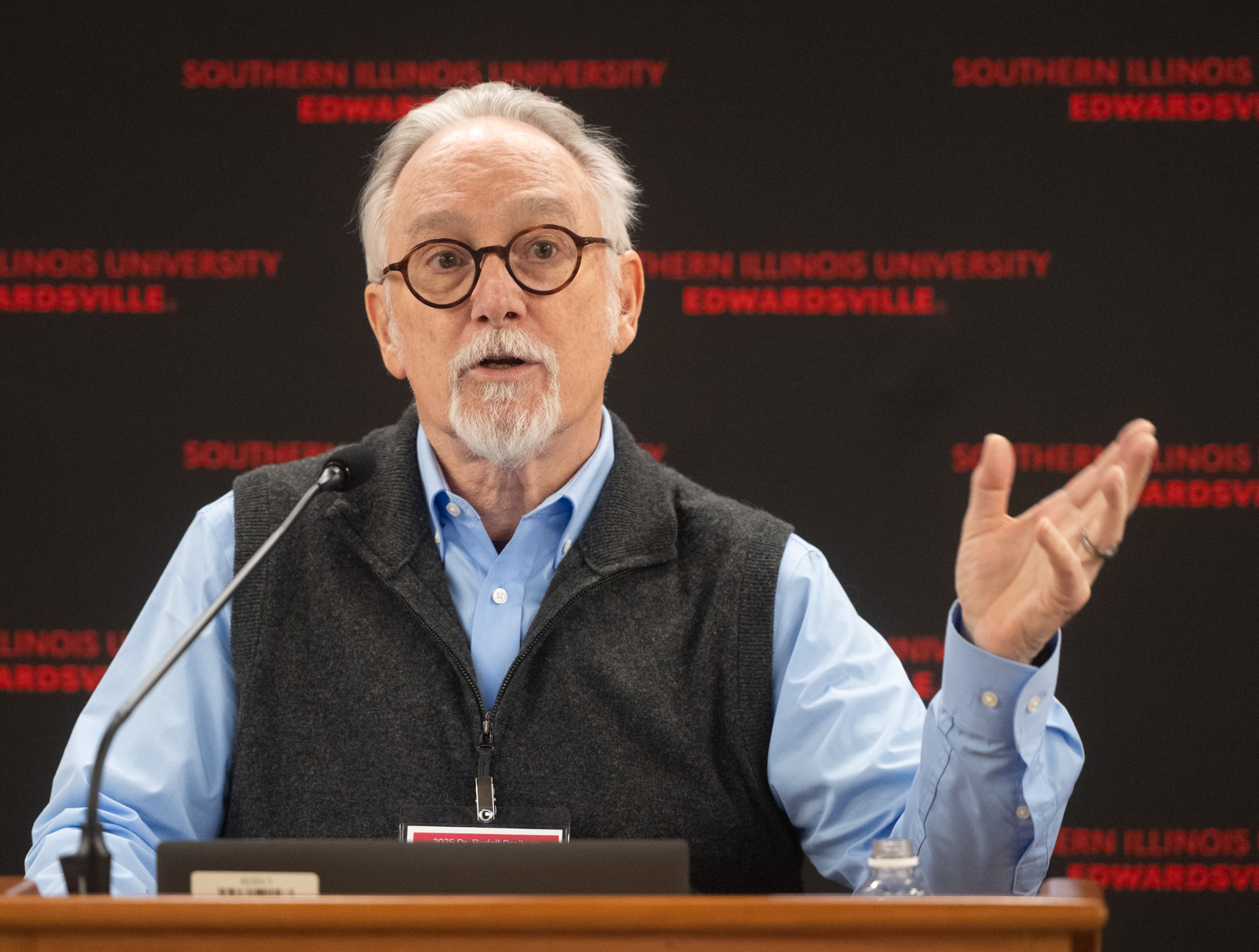 photo of Rocky Garrison speaking at a lectern against a black SIUE branded background, gesturing with his right hand