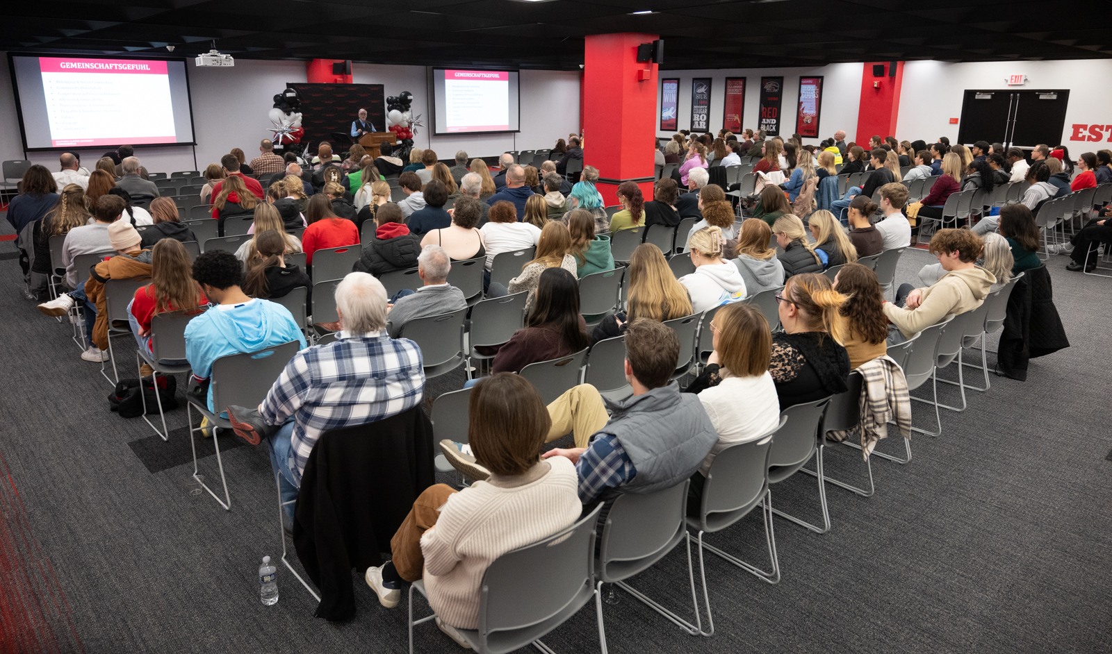 Photo of the Legacy Room with a substantial amount of people seated in chairs to listen to Dr. Garrison's lecture. There are slides on the walls outlining his keynote displayed behind him.