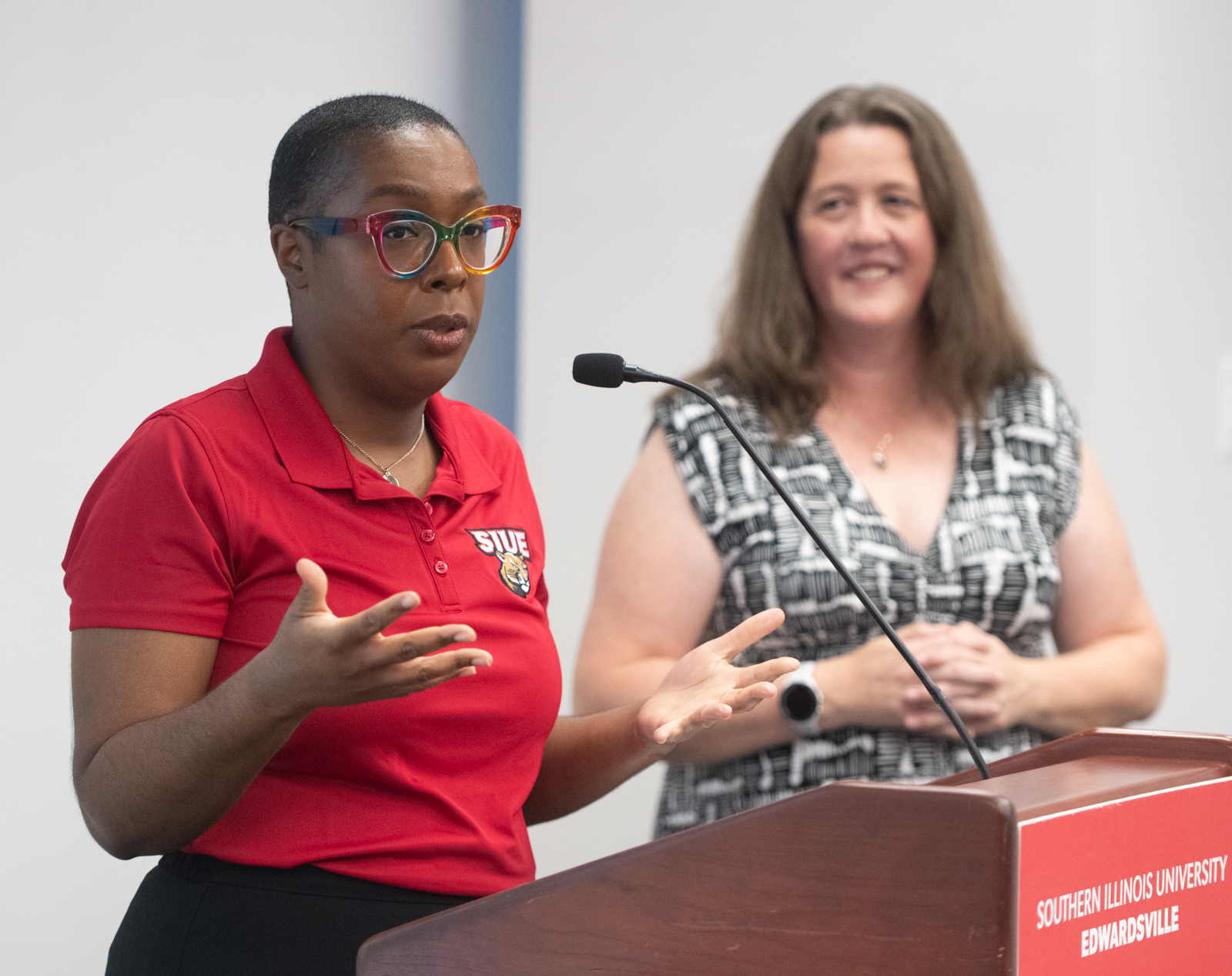 “A woman in a red SIUE polo shirt speaks at a podium with a microphone, gesturing with her hands as she addresses an audience. Another woman stands smiling in the background.”
