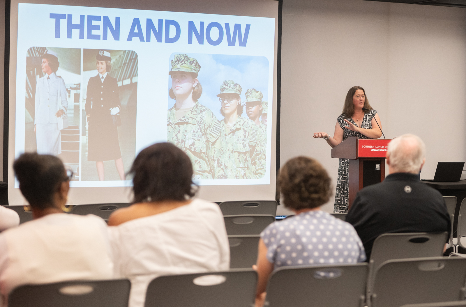 “A woman stands at a podium speaking to an audience seated in rows of chairs. Behind her, a large projected slide reads ‘Then and Now’ and shows historical and modern images of women in military uniforms.”