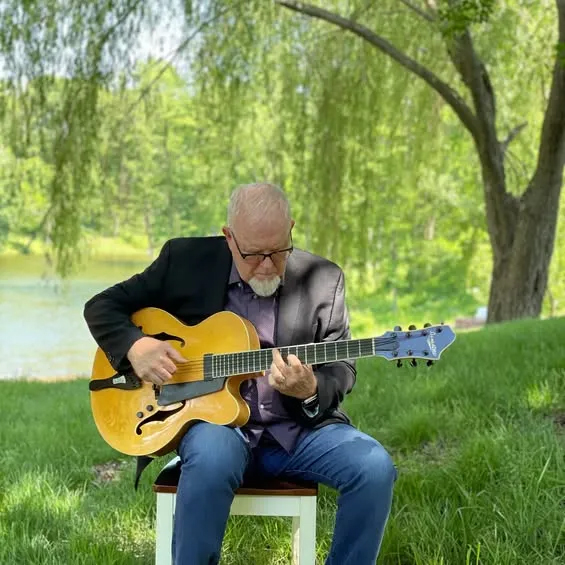 Rick Haydon sits outdoors playing a semi-hollow guitar beneath a weeping willow near a small pond.