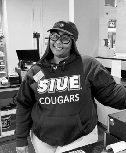A person wearing an SIUE Cougars hoodie and cap smiles while standing behind a counter indoors.