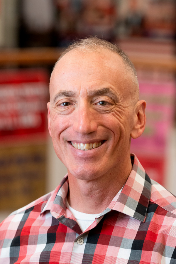 Professional portrait of Tom Spudich smiling and wearing a red plaid shirt against a softly blurred indoor background.