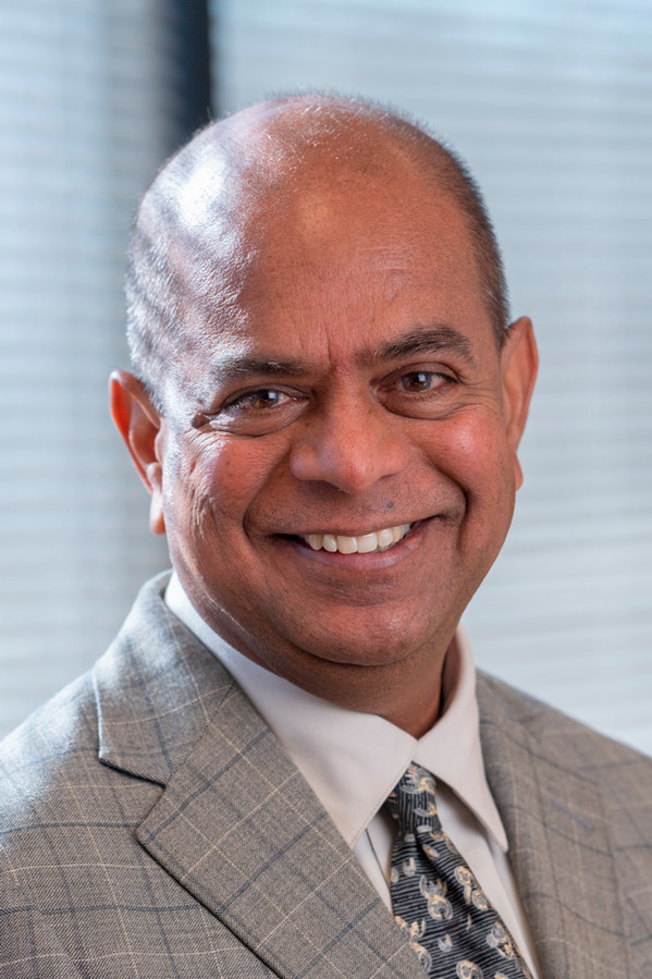 Professional headshot of Vijay Singh Vadlani smiling in a light gray suit and patterned tie against a softly lit neutral background.