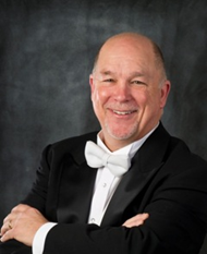 Studio portrait of a smiling man in a black tuxedo with a white bow tie, posed with arms crossed against a dark mottled backdrop.