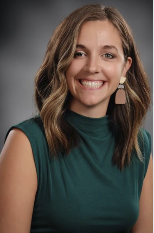 Professional headshot of Hiller with shoulder-length wavy hair wearing a sleeveless green top and geometric earrings against a gray studio background.