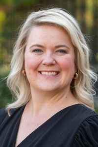 Portrait of Mindy Dilley outside wearing delicate hoop earrings, a black blouse and a smile