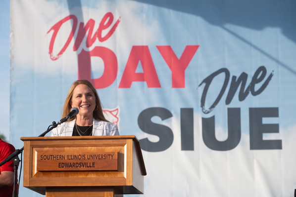 Lisa Spencer smiles while speaking at a wooden podium labeled “Southern Illinois University Edwardsville” during the One Day, One SIUE celebration, with a large “One Day, One SIUE” banner in the background.
