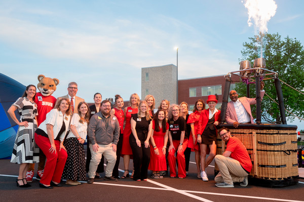 A dozen staff members in University Advancement line up in front of a half inflated hot air balloon while Chancellor Minor stands in the basket activating the propane ignition creating a bright glow in the parking lot in front of the Health sciences