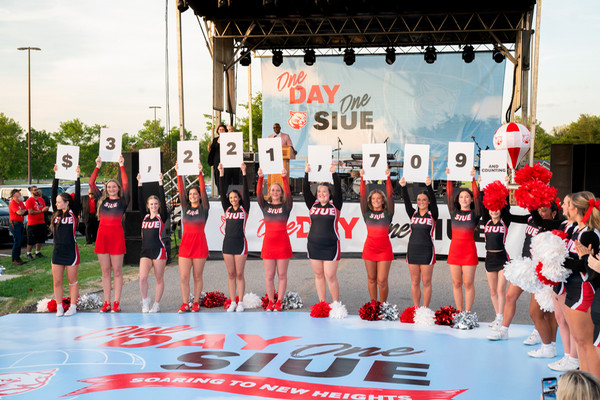 SIUE Cheerleaders and dance team members wearing uniforms and sporting pop poms line up on the One Day One SIUE dance floor in front of a large outdoor stage holding up numbers that say 3,221,709