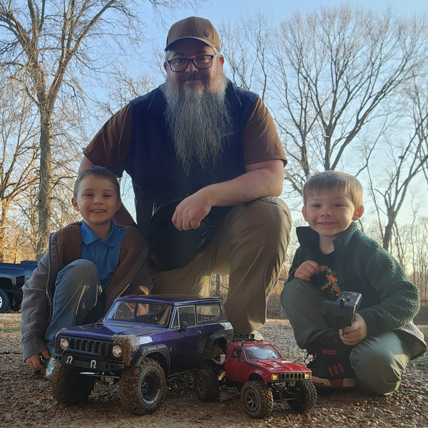 Eric Barbee kneels outdoors with his two young sons beside remote-controlled off-road vehicles. The family smiles for a photo on a gravel path surrounded by leafless trees on a clear day.