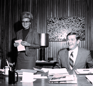 Black and white photo of Wilma Jene wearing a dress suit and a well-groomed beehive style updo standing beside President Rendleman at his desk. 