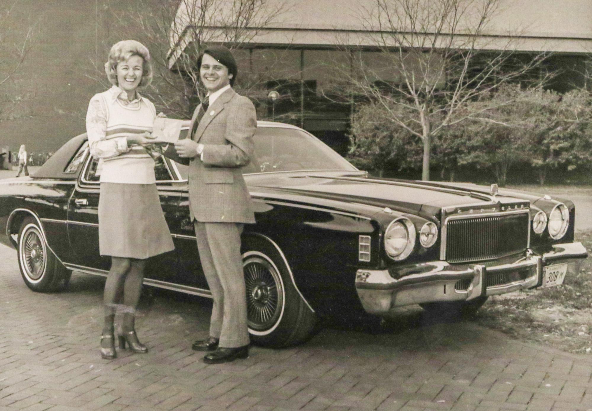 Black-and-white archival photo of Wilma Jene Bond and a gentleman in a suit smiling beside a vintage car parked on the quad outside SIUE's Dunham Hall.