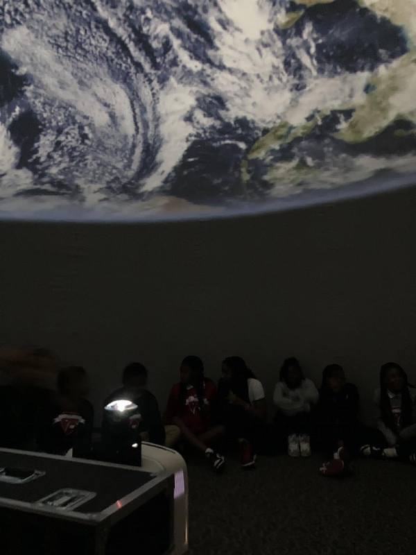 Inside a dark planetarium, a group of students sits along the curved wall beneath a projection of Earth’s swirling clouds and continents. A star projector glows in the foreground, casting faint light across the room.