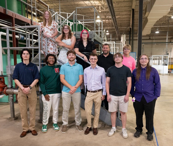 Group photo of twelve new graduates in the ERTC building where water is processed. They are standing on one of the staircases supporting the laboratory infrastructure and are smiling