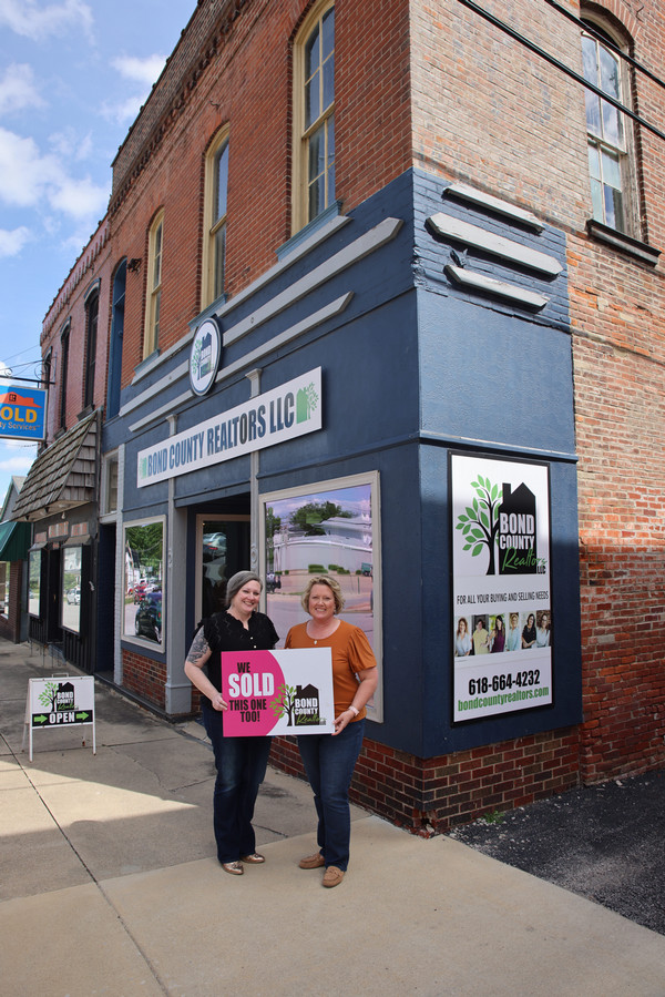 Two realtors standing outside exterior of realty office holding SOLD sign