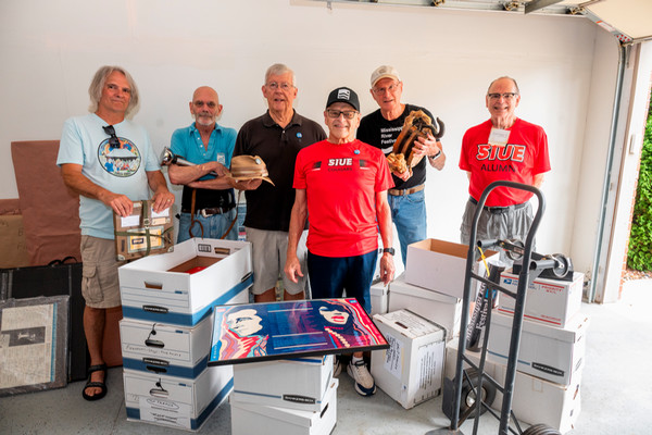 Lyle Ward stands in his garage amongst fellow MRF workers surrounded by cardboard boxes filled with concert posters. The six men stand together holding artifacts like a rope pulley, a tent pole finial, and a hat. Lyle wears an SIUE shirt.
