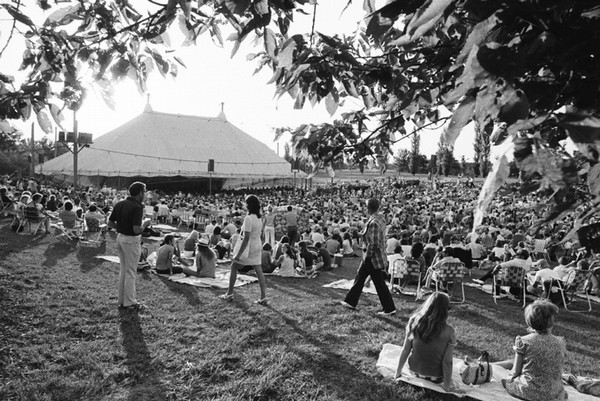 An archival photo of the historic Mississippi River Festival in 1969. It is black and white and shows a vast lawn filled to the brim with people in lawn chairs surrounding a stately white tent surrounded by greenery and foliage.
