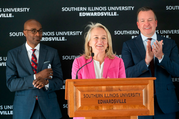 U.S. Rep. Nikki Budzinski smiles at an SIUE podium, with Chancellor James T. Minor to her left and Deputy Governor Andy Manar applauding to her right during the NCERC expansion announcement.